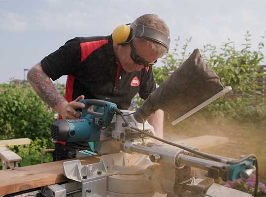 Håndverker i svart og rødt sager tre ute med blå kappsag og gult hørselsvern