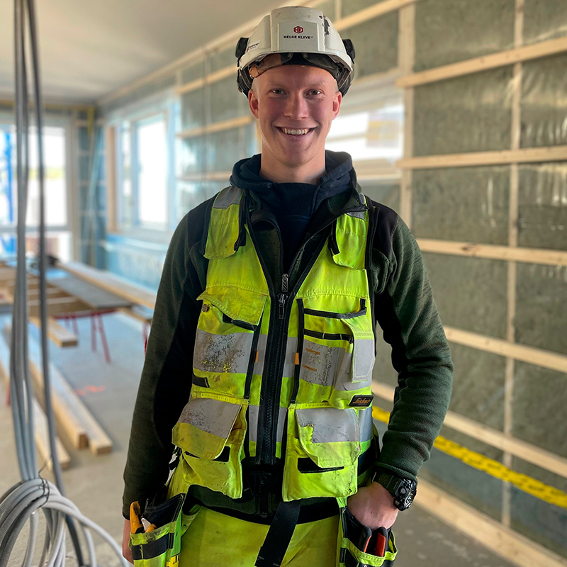 Bygningsarbeider i gul refleksvest og hvit hjelm i uferdig rom med synlige stendere