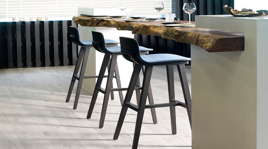 Moderne bardisk med ujevn treplate, tre blågrå barkrakker og vinglass ved vindu