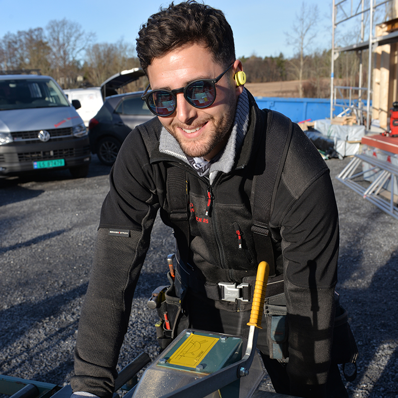 Håndverker i svart arbeidstøy med gult hørselvern og verktøybelte ved sagbord på byggeplass