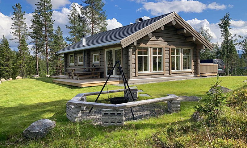 Grå tømmerhytte i sol, stor grønn plen med trekantet bålsted og furuskog bak.