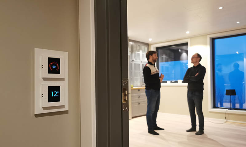 To digitale termostater (rød/blå) på gangvegg, to menn snakker i lyst parkettrom med vindu