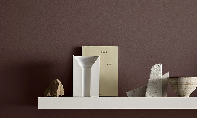 Hvit hylle mot plommefarget vegg med abstrakte hvite/beige skulpturer og lysegul bok.