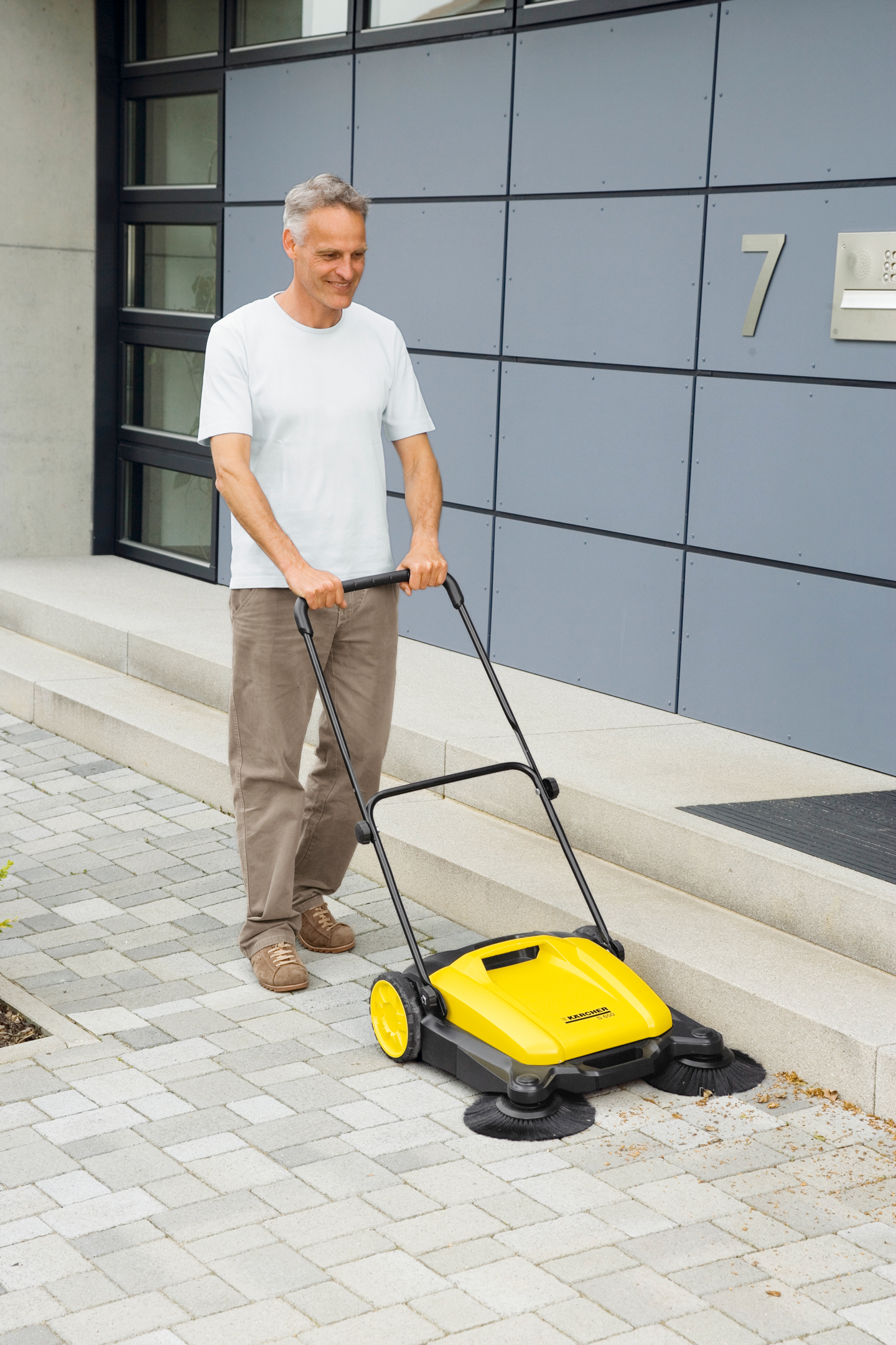 Mann i hvit t-skjorte skyver gul feiemaskin på grå brostein ved moderne hus.