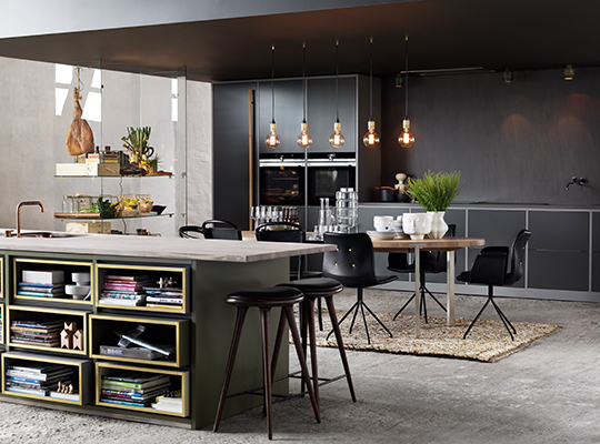 Moderne kjøkken i grått og grønt med lys steinbenk, hylle med bøker, svarte stoler og hengelamper.