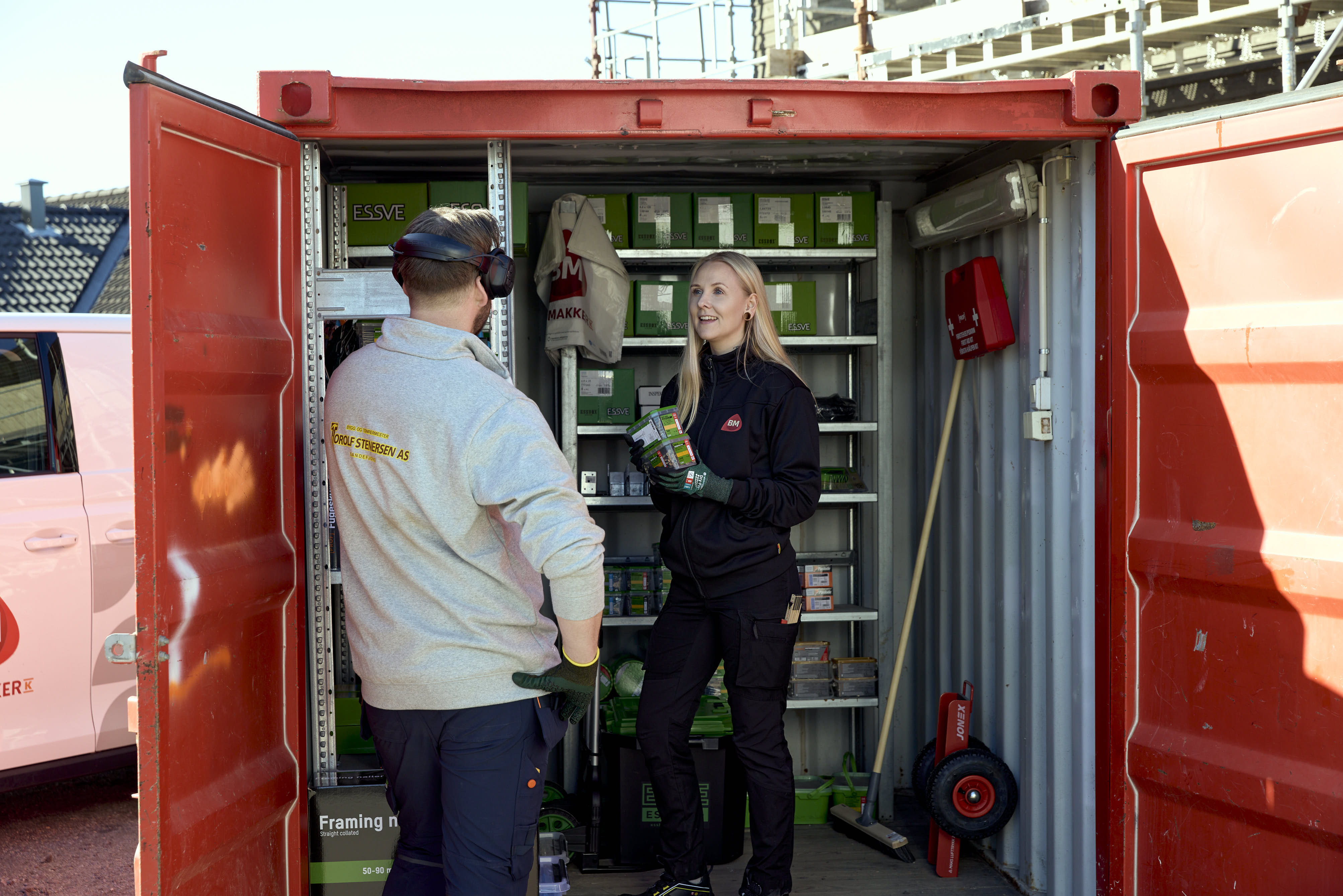To håndverkere i åpen rød container med grønne ESSVE-kasser på byggeplass.