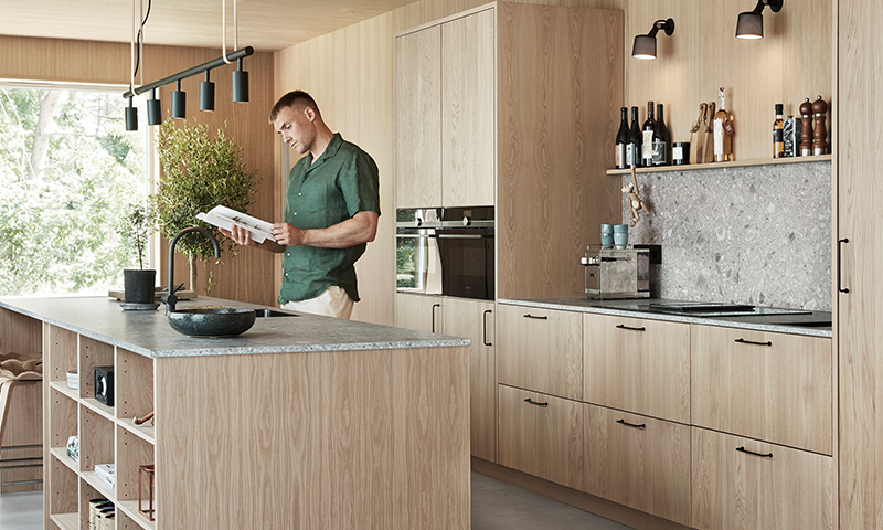 Lyst kjøkken i tre med grå steinbenk, åpen kjøkkenøy, hengelamper og person som leser bok.