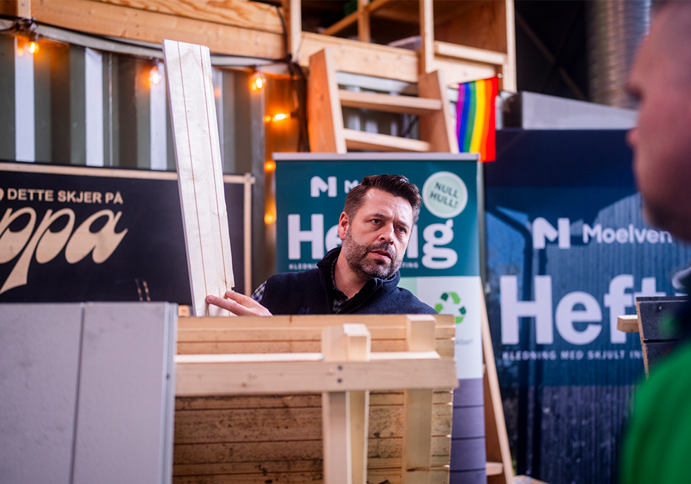 Mann holder treplanker ved en Moelven-stand med skilt og regnbueflagg i bakgrunnen.