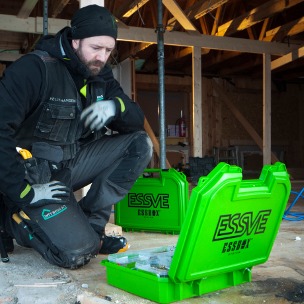 Bygningsarbeider i uferdig trehus, kneler ved gulvet med åpne, grønne ESSVE-kasser.