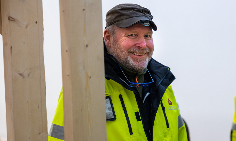 Person i neongul refleksjakke og grå caps foran trebjelker på byggeplass