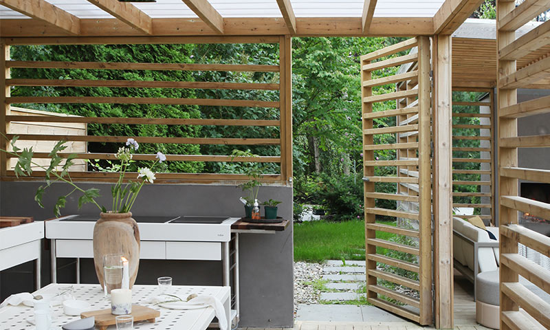 Utendørs treglassert pergola med hvitt kjøkkenbord, blomster i vase, grønn hage utenfor.