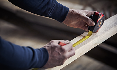 Hender som måler en treplanke med svart-rødt målebånd og gul stripe
