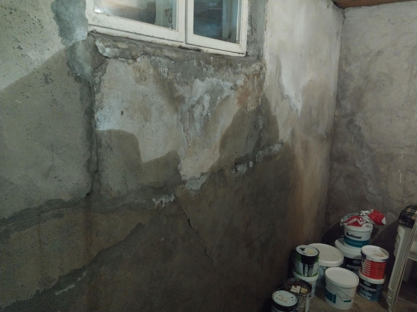 Grå, fuktflekkete kjellervegg med sprekk under vindu og stabler av malingsspann på gulvet