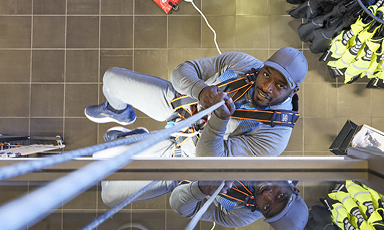 Arbeider i grå klær og blå hjelm klatrer i oransje sele og tau over grått flisgulv ved gule vester.