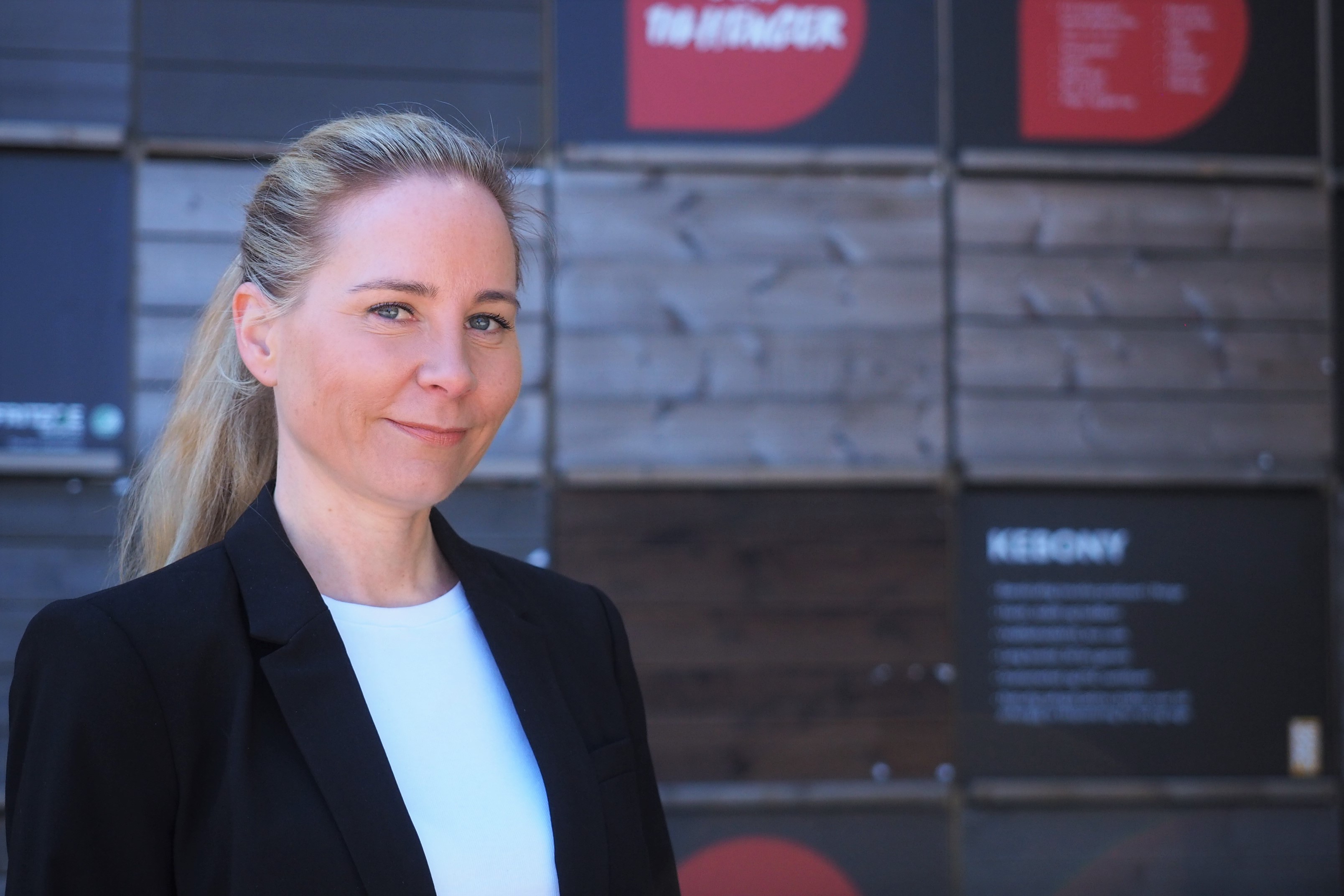 Blond person i svart blazer og hvit skjorte foran trefarget vegg med røde og svarte skilt.