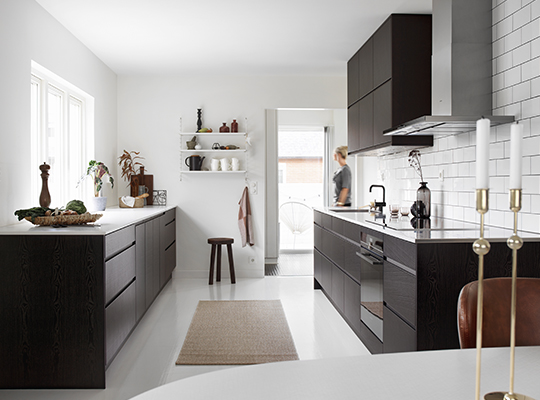 Lyst minimalistisk kjøkken med mørke skap, hvite fliser, beige løper og kvinne i bakgrunnen