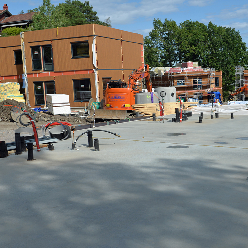 Byggeplass med brunt modulbygg under oppføring, oransje gravemaskin, rør og kabelruller.