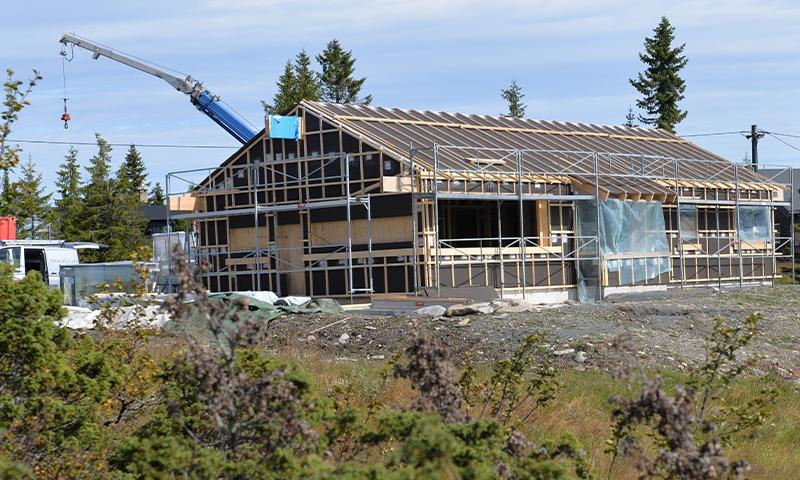Trehus under oppføring med stillas og kran, omgitt av skog under blå himmel.