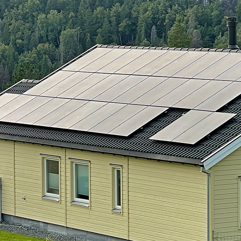 Gult enetasjes hus med svarte solcellepaneler på tak og grønne skogstrær i bakgrunnen.