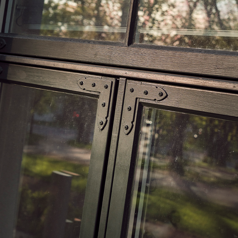 Nærbilde av mørkegrønn vindusramme med jernbeslag og glass som speiler grønt uteområde