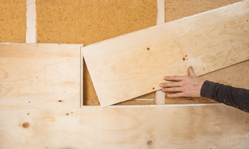 Hånd plasserer lyst kryssfinerpanel foran brunt isolasjonsmateriale på vegg.