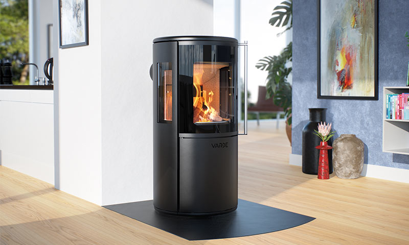 Svart sylindrisk Varde-vedovn med glødende flammer i lyst, moderne stue med kunst og planter.