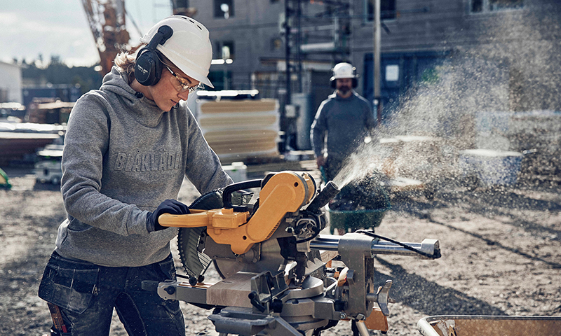 En arbeider med hvit hjelm og hørselsvern bruker gul kapp- og gjæringssag på byggeplass