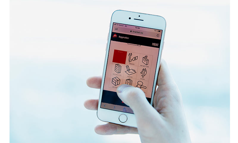 Hånd holder hvit telefon som viser Byggmakker-nettside med røde og sorte ikoner på lys bakgrunn.