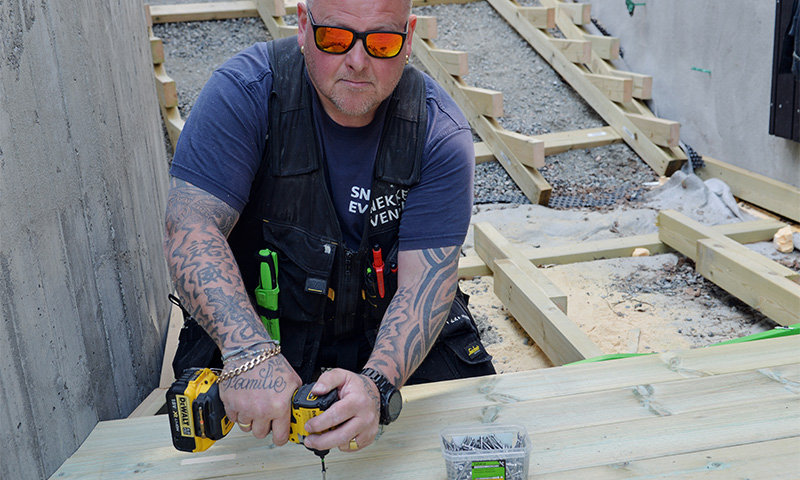 Tattooert håndverker skrur lyst tredekke med gul drill på byggeplass.