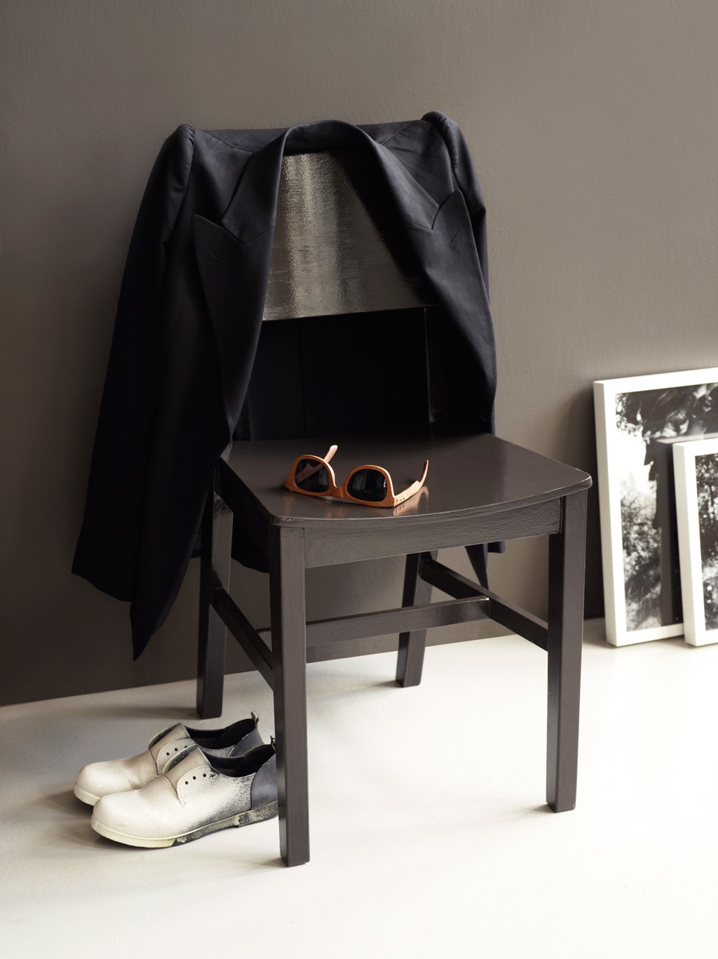 Svart stol mot grå vegg med mørk jakke over rygg, brune solbriller på sete og hvite sko ved siden.