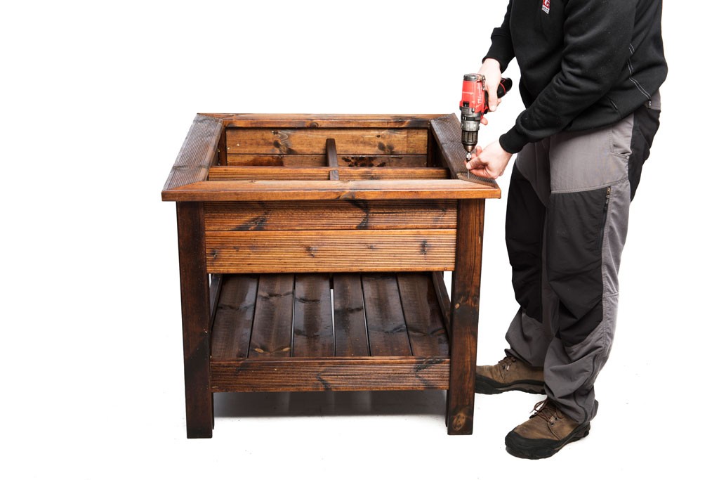 Person i arbeidsklær med bor skrur sammen mørk brun trebenk med hylle på hvit bakgrunn