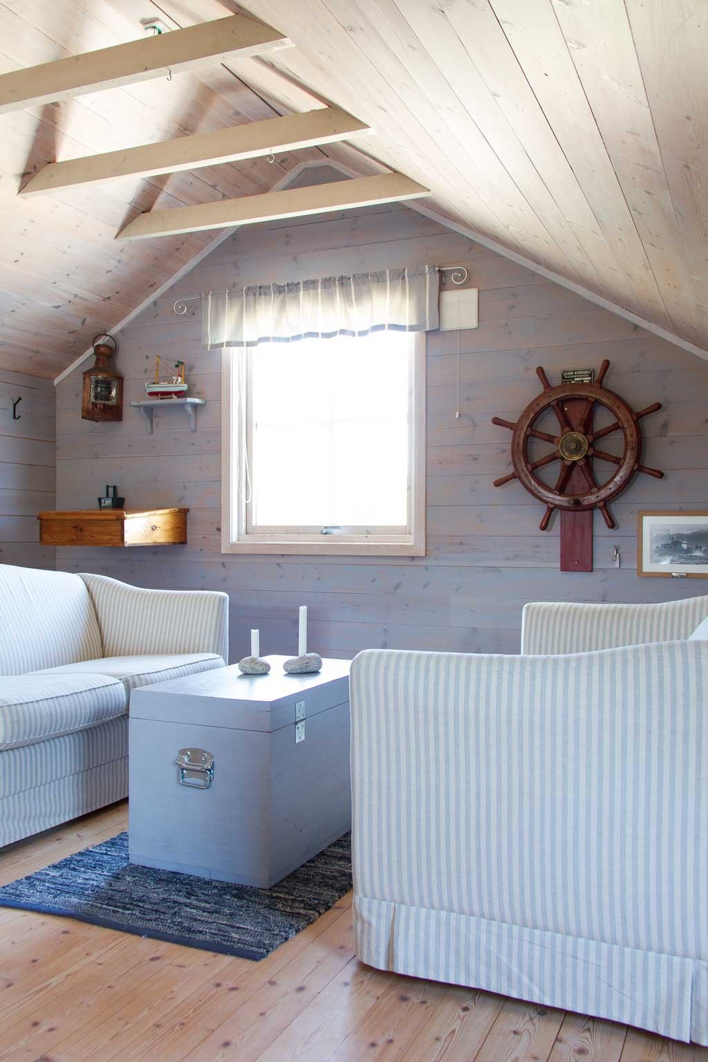 Lyst loftstue med lysegrå stripete sofaer, grå kistebord, stearinlys og skipshjul på trepanel.