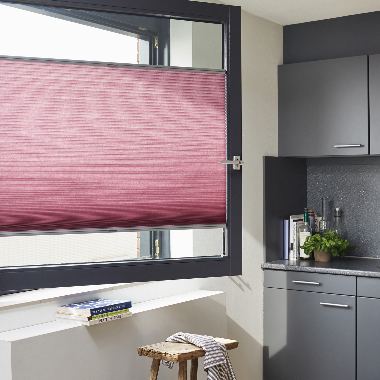 Kjøkken med åpent vindu og rosa plisségardin, grå skap, trekrakk med klut.