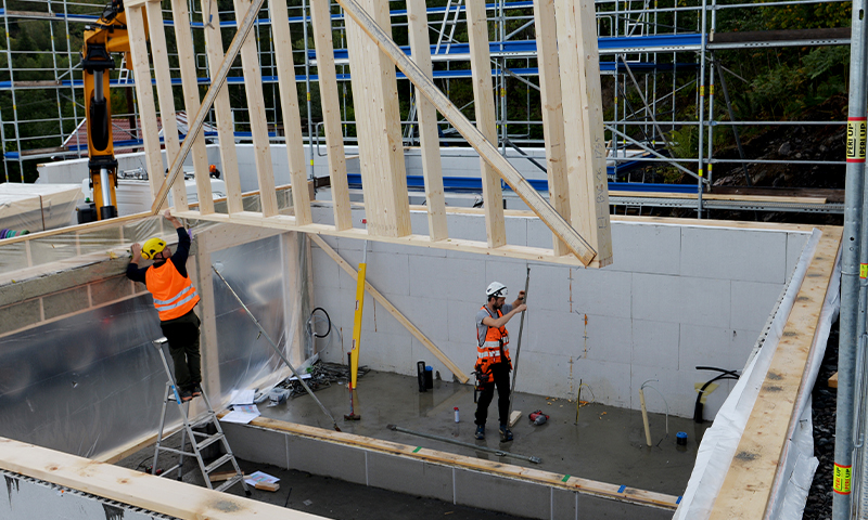 To arbeidere i oransje vester og hvite hjelmer løfter prefabrikert veggstamme på byggeplass