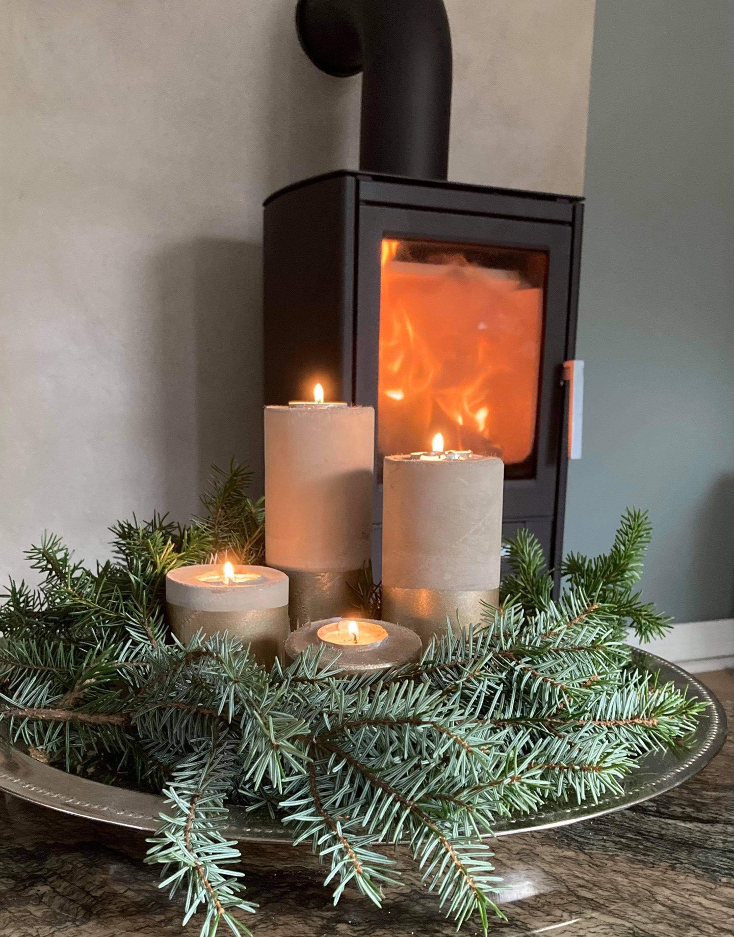 Flammer i svart vedovn bak sølvfat med fire grå adventslys i grønn furukrans