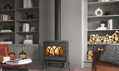 Koselig stue med svart vedovn i midten, grå hyller med vedstokker, bøker og dekor.