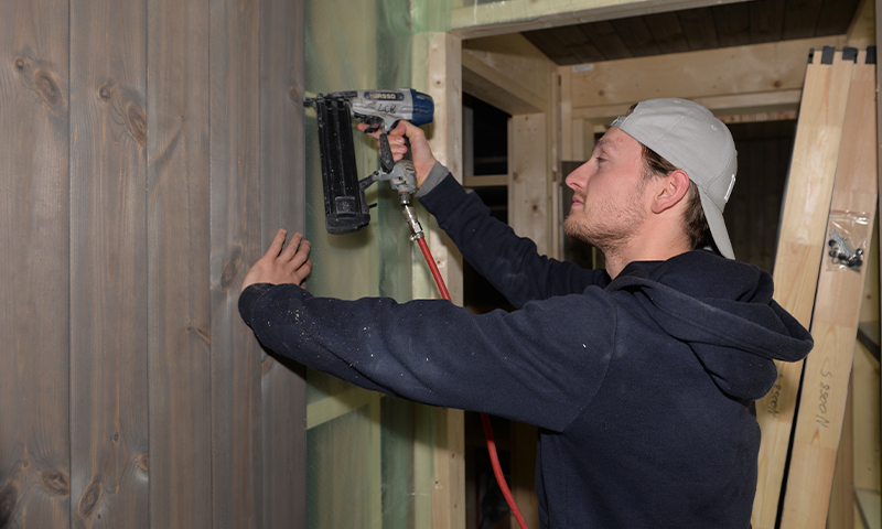 Håndverker i mørk genser og lys cap fester gråbeiset panel i uoppusset trevegg med spikerpistol.
