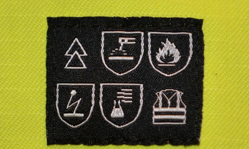 Svart stoffmerke med seks hvite sikkerhetspiktogrammer mot gul sikkerhetsvest