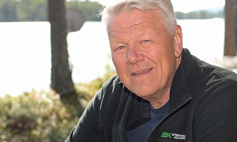Mann i svart fleecejakke med grønn logo ved vannkant, trær og sollys i bakgrunnen.