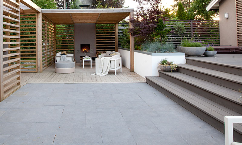 Moderne uteplass med grå fliser, trekledning, pergola med peis, hvite sittegrupper og grønne planter