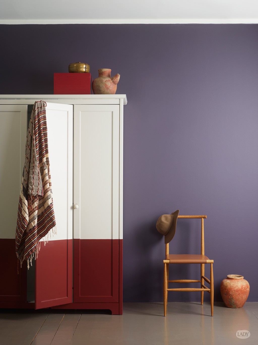 Hvit-rødt skap mot lilla vegg, stripet tekstil, trestol med hatt og terrakottapotter.