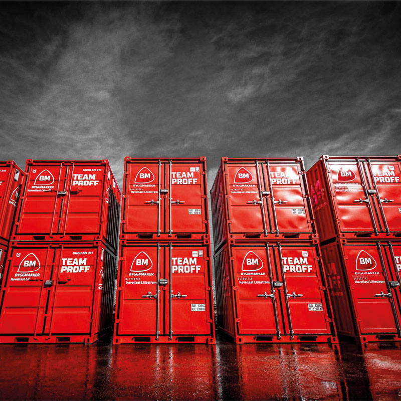 Røde BM Team Proff byggecontainere stablet på våt asfalt under dramatisk skyet himmel