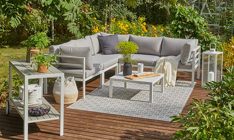 Moderne uteområde med L-formet grå sofa, hvitt salongbord, grønne planter og gule blomster