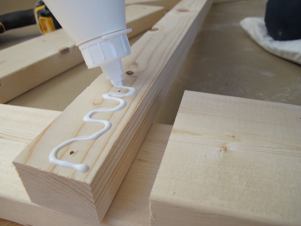 Flaske med hvit lim påfører bølget stripe på lyst furubord, verktøy i bakgrunnen