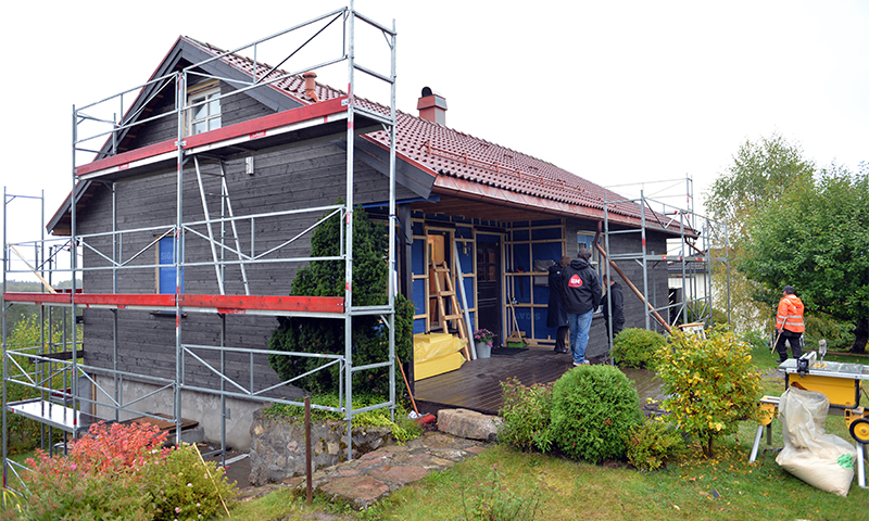 Grått trehus med rødt tak og stillas, håndverkere jobber i grønn hage.
