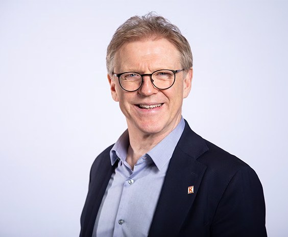 Mann iført mørk blazer og lyseblå skjorte mot lys bakgrunn med partilogo på jakkeslaget.