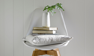 Hvit stol med grå pute, bokstabel og hvit vase med grønne blader foran hvit panelvegg.