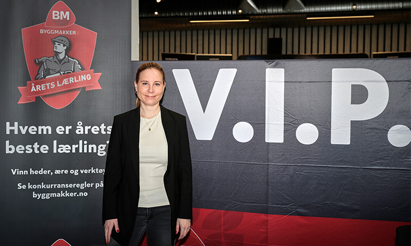 Person i svart blazer og hvit genser foran rødt og sort Byggmakker Årets lærling-banner og VIP-skilt.