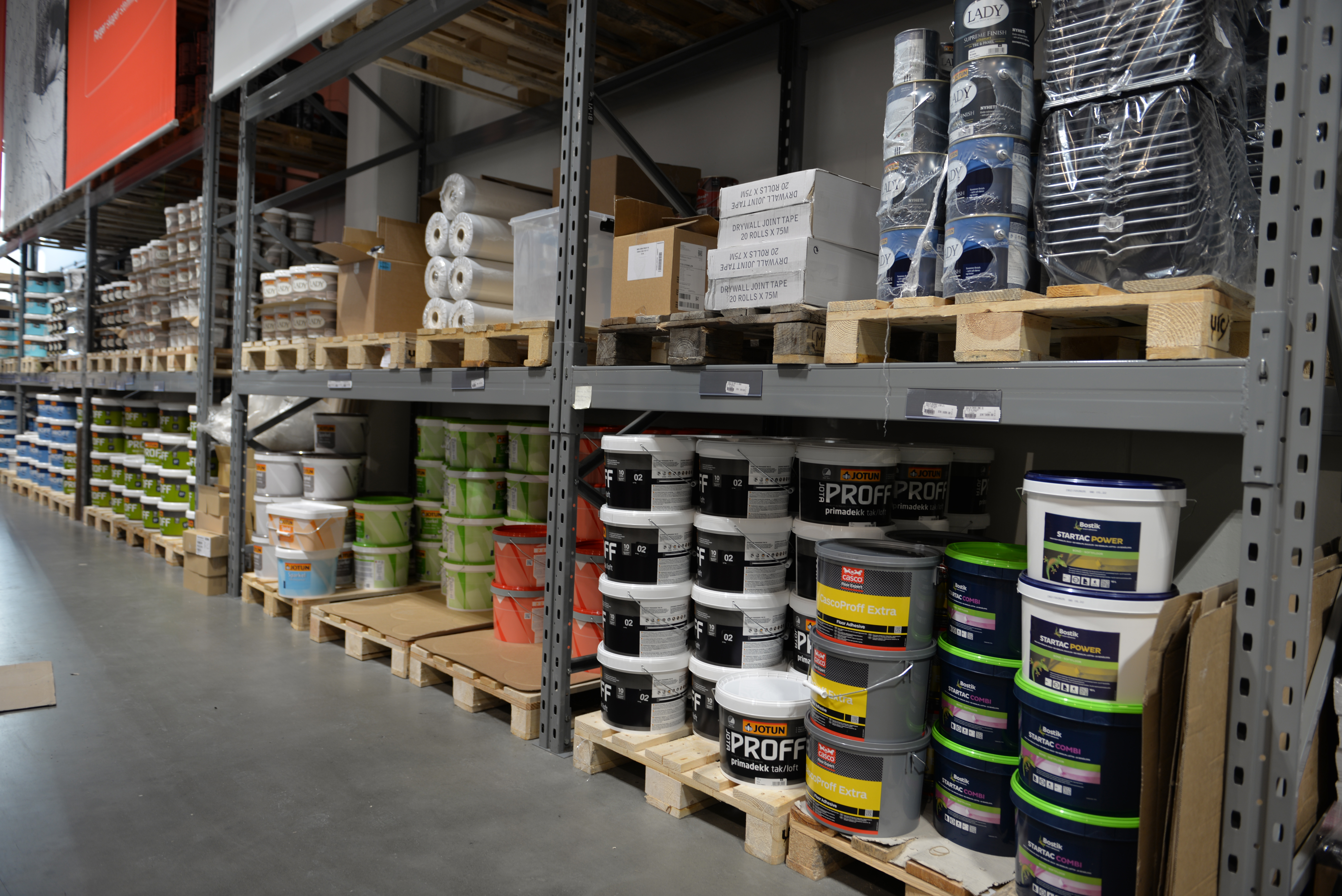 Stålhylle i byggevarehus med fargerike malingsspann, tapebokser og ruller på trepaller.