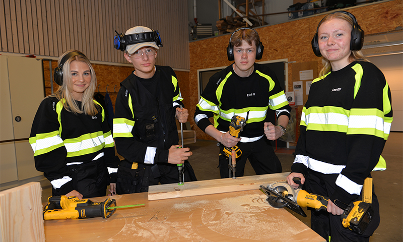 Fire arbeidere i svart arbeidsdress med gule refleksstriper i treverksted med elverktøy og treplanker